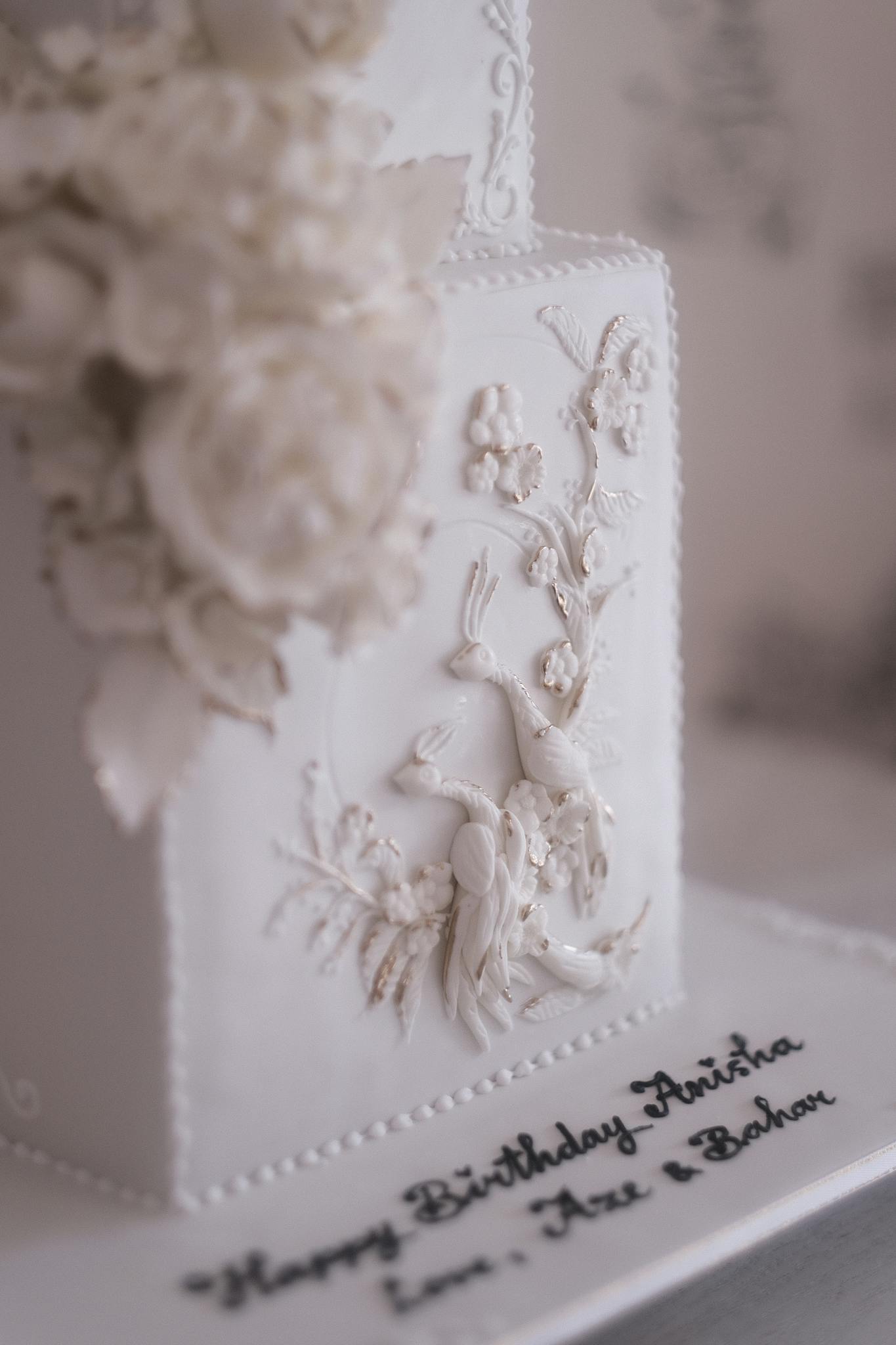Close-up of an intricate white floral design on a square wedding cake, with birthday wishes.
